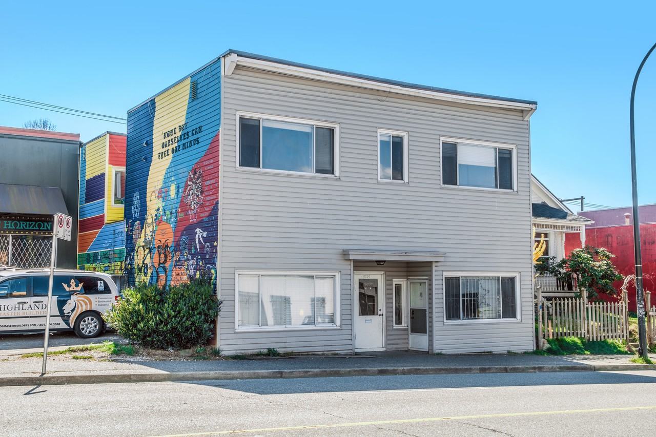 A two-story building with a colorful mural on one side, located on a street with a parked vehicle and greenery in front.
